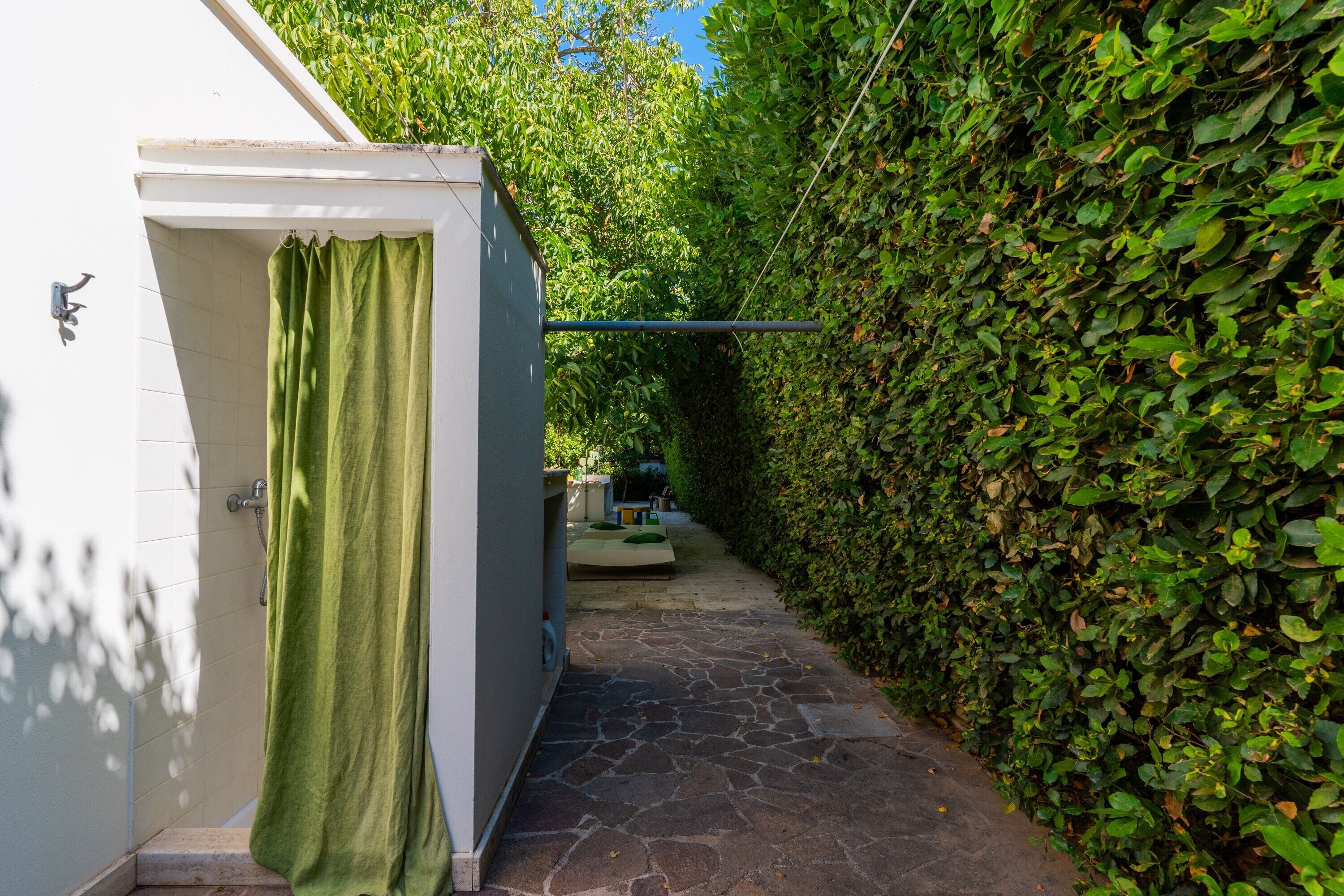 A narrow stone pathway leading to a white building with a green curtain, surrounded by dense green foliage.