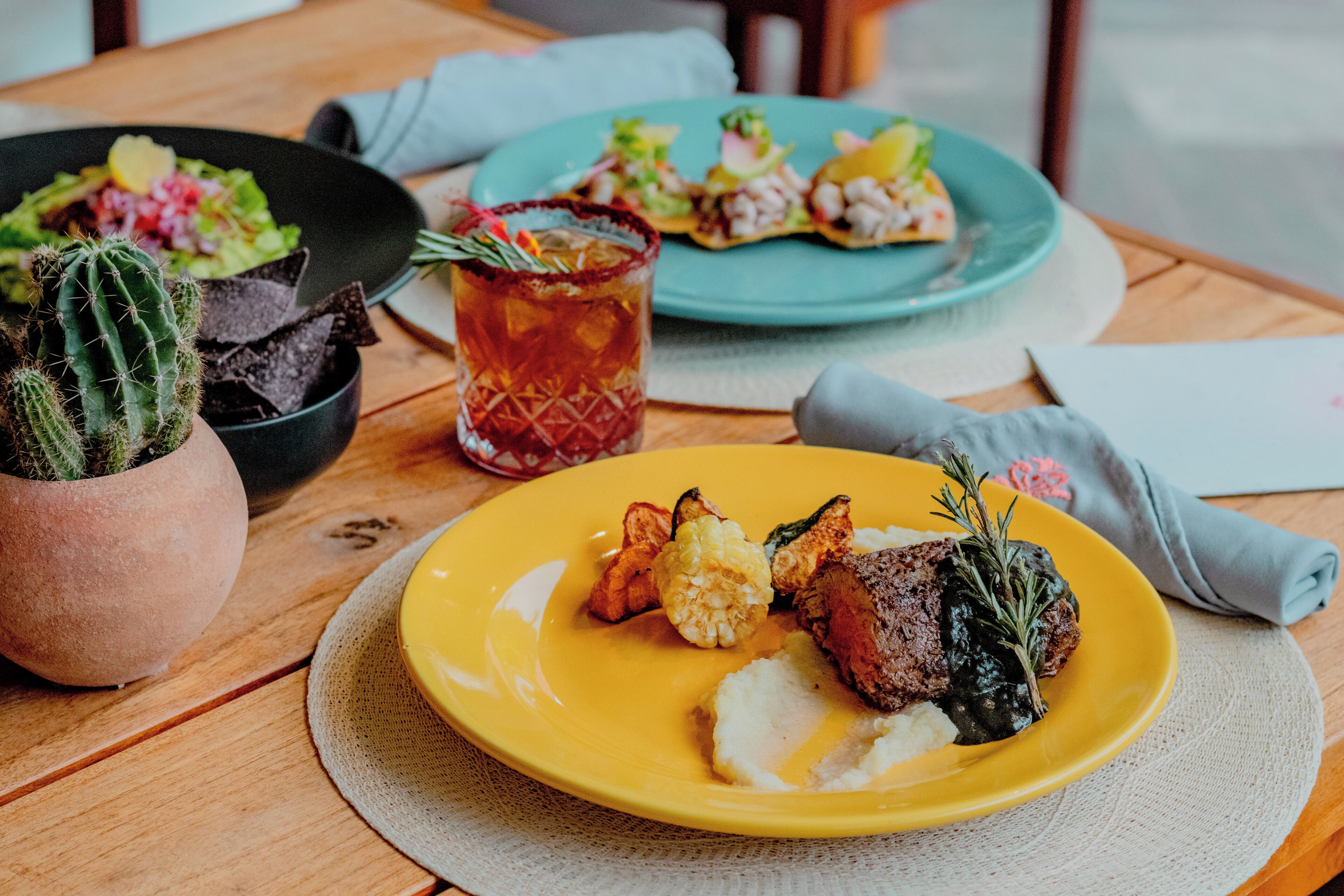 A dining table with a yellow plate of food, a green plate with appetizers, a glass of iced tea, and a cactus plant.