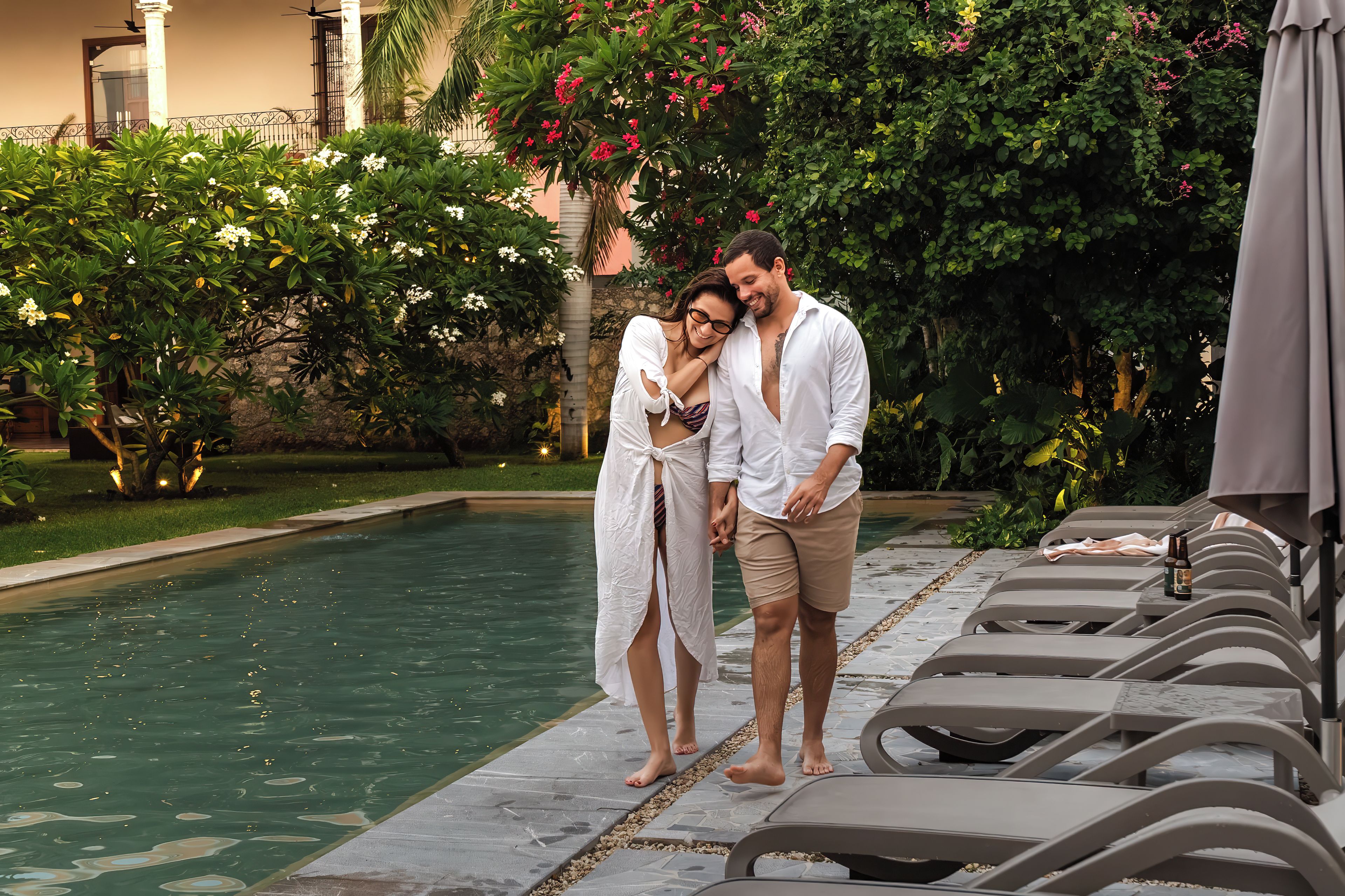 A couple standing by a pool with lounge chairs and a garden in the background.