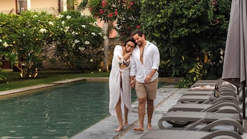 A couple standing by a pool with lounge chairs and a garden in the background.