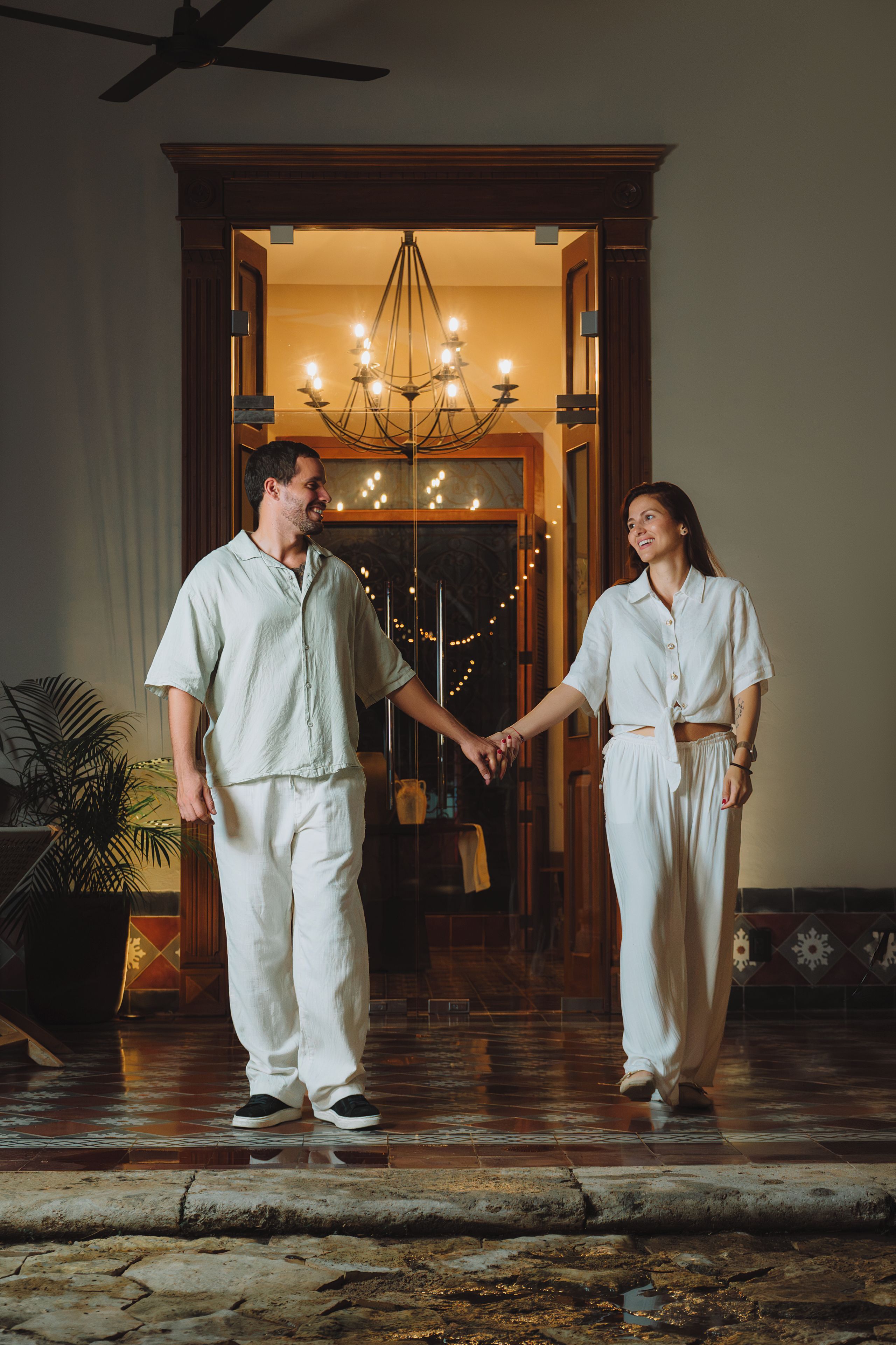 A couple holding hands in a warmly lit foyer with a chandelier and a potted plant.