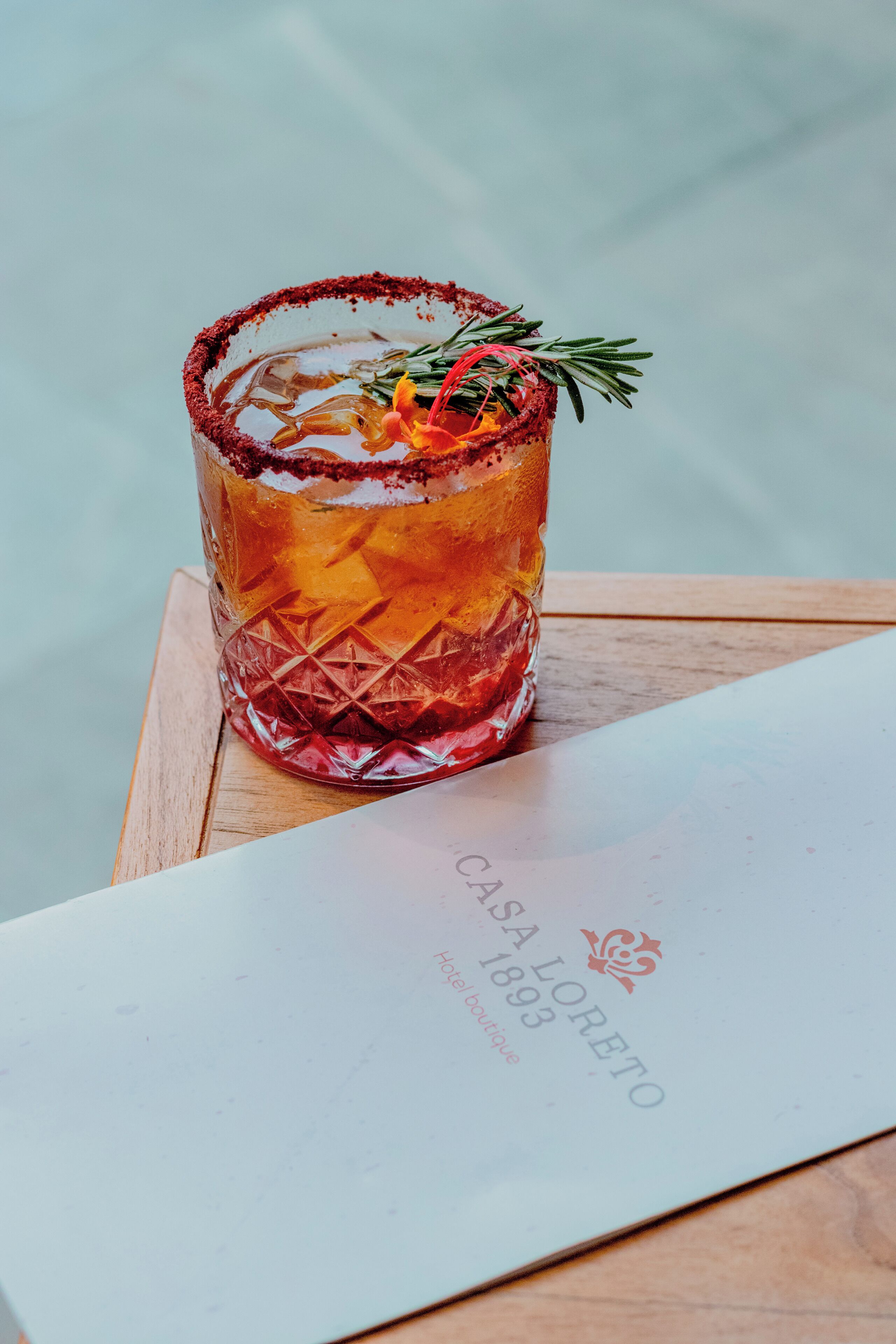 A glass of amber-colored drink with a rim of red salt, garnished with a sprig of rosemary, on a wooden table with a menu.