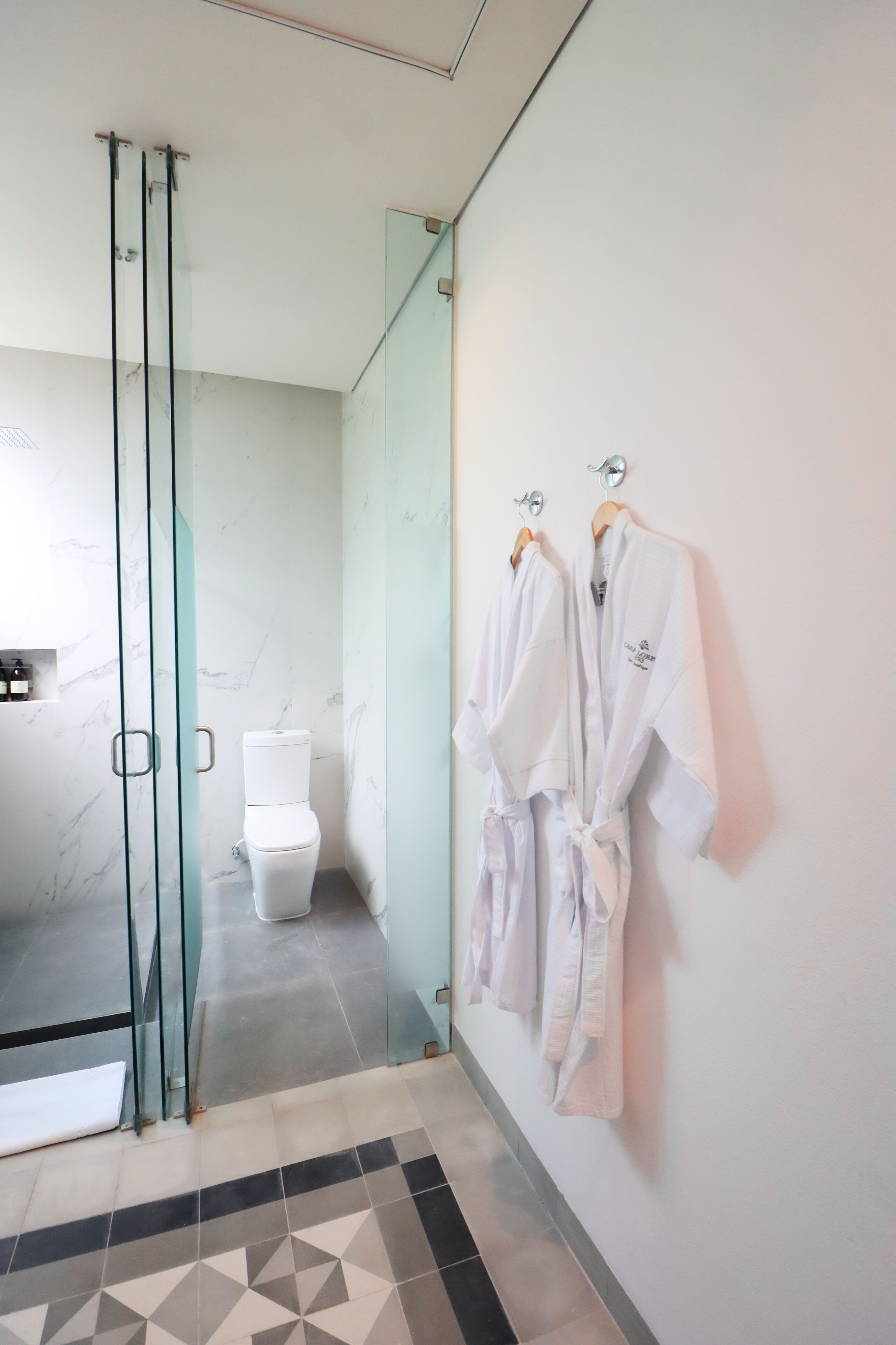Ein modernes Badezimmer mit Duschkabine aus Glas, einer Toilette und zwei weißen Bademänteln, die an einer Wand hängen.