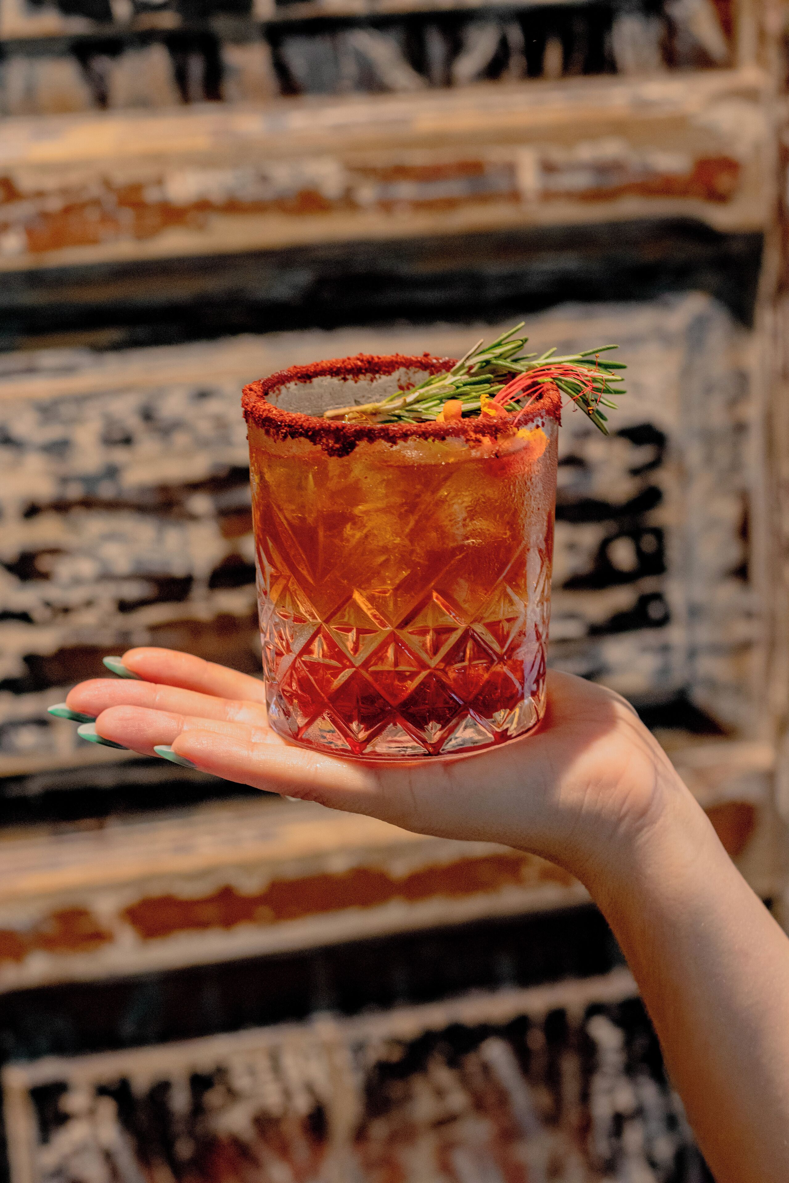 A hand holding a textured glass with a garnish, against a patterned wall.