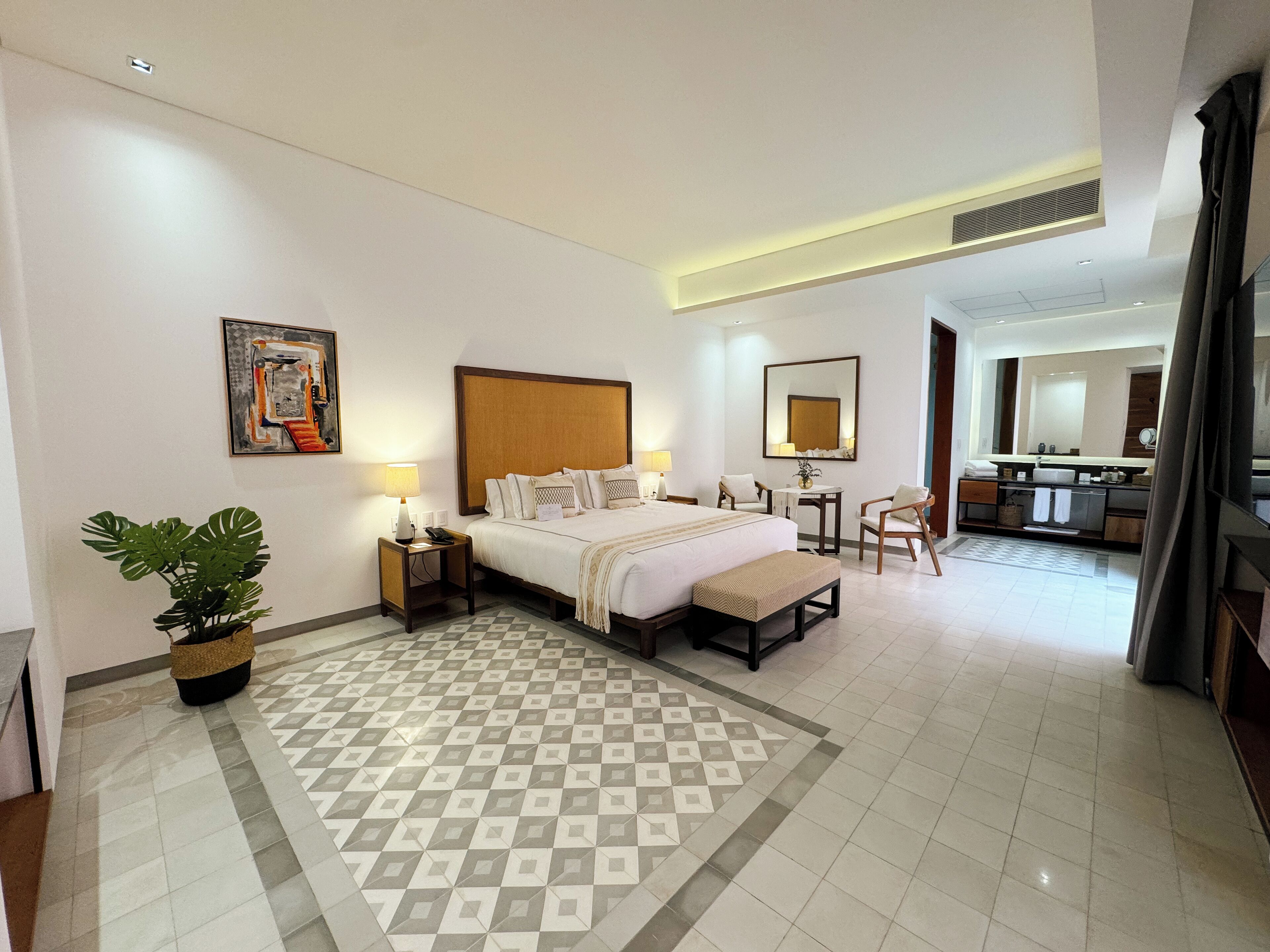 A hotel room with a large bed, bedside tables, a desk, a chair, a potted plant, and a bathroom visible through an open door.