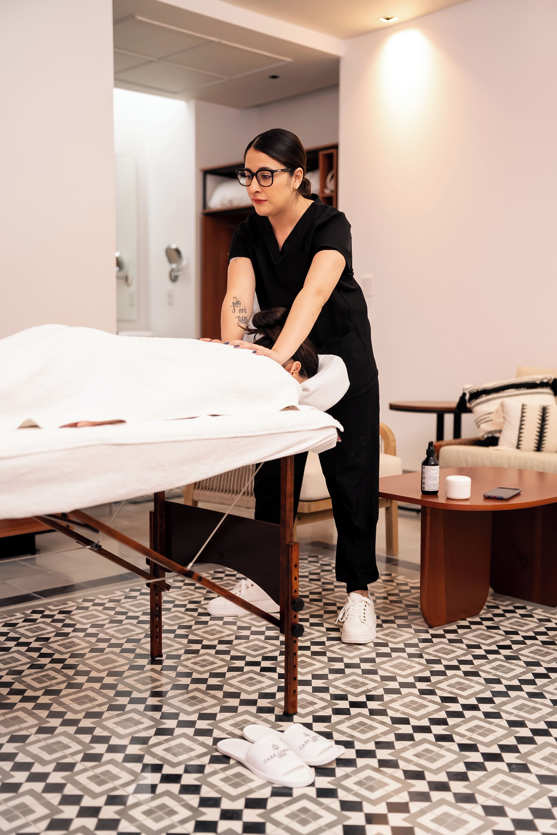A person in black attire preparing a bed in a room with patterned floor tiles and a wooden table with a bottle and a cup.