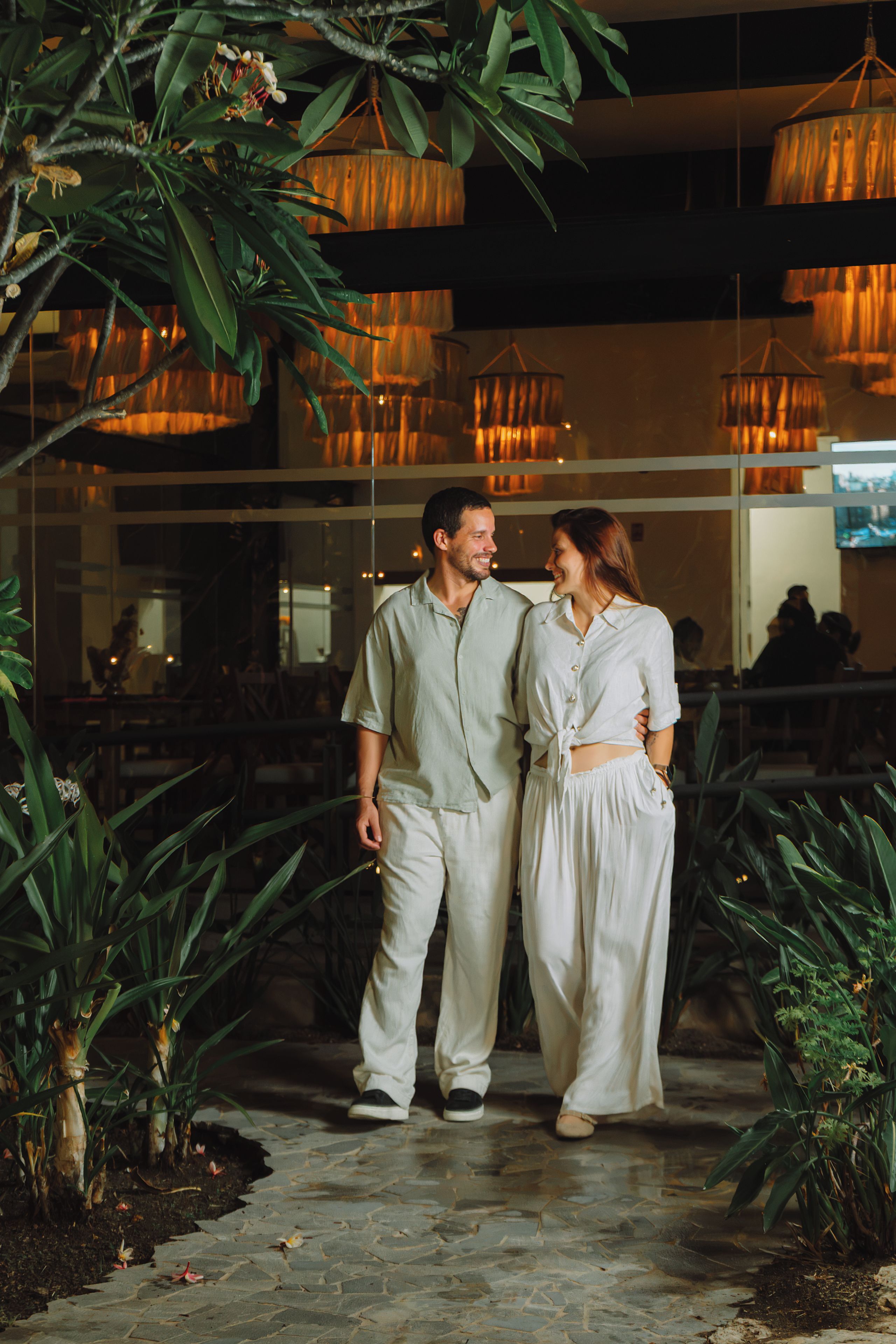 A couple standing in a well-lit indoor area with hanging lamps and potted plants.