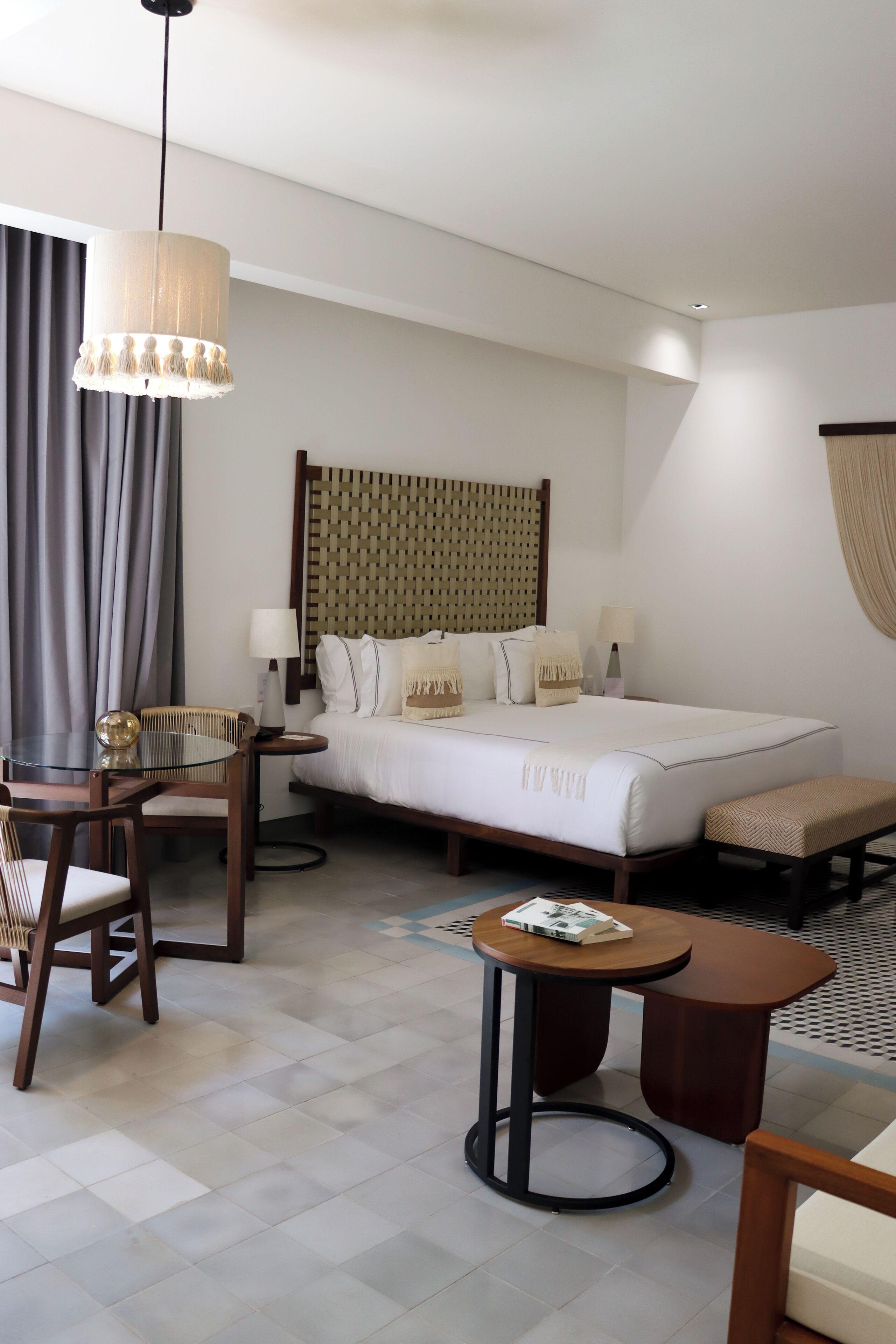 A hotel room with a bed, bedside tables, a round table, a chair, a hanging light fixture, and a patterned wall headboard.
