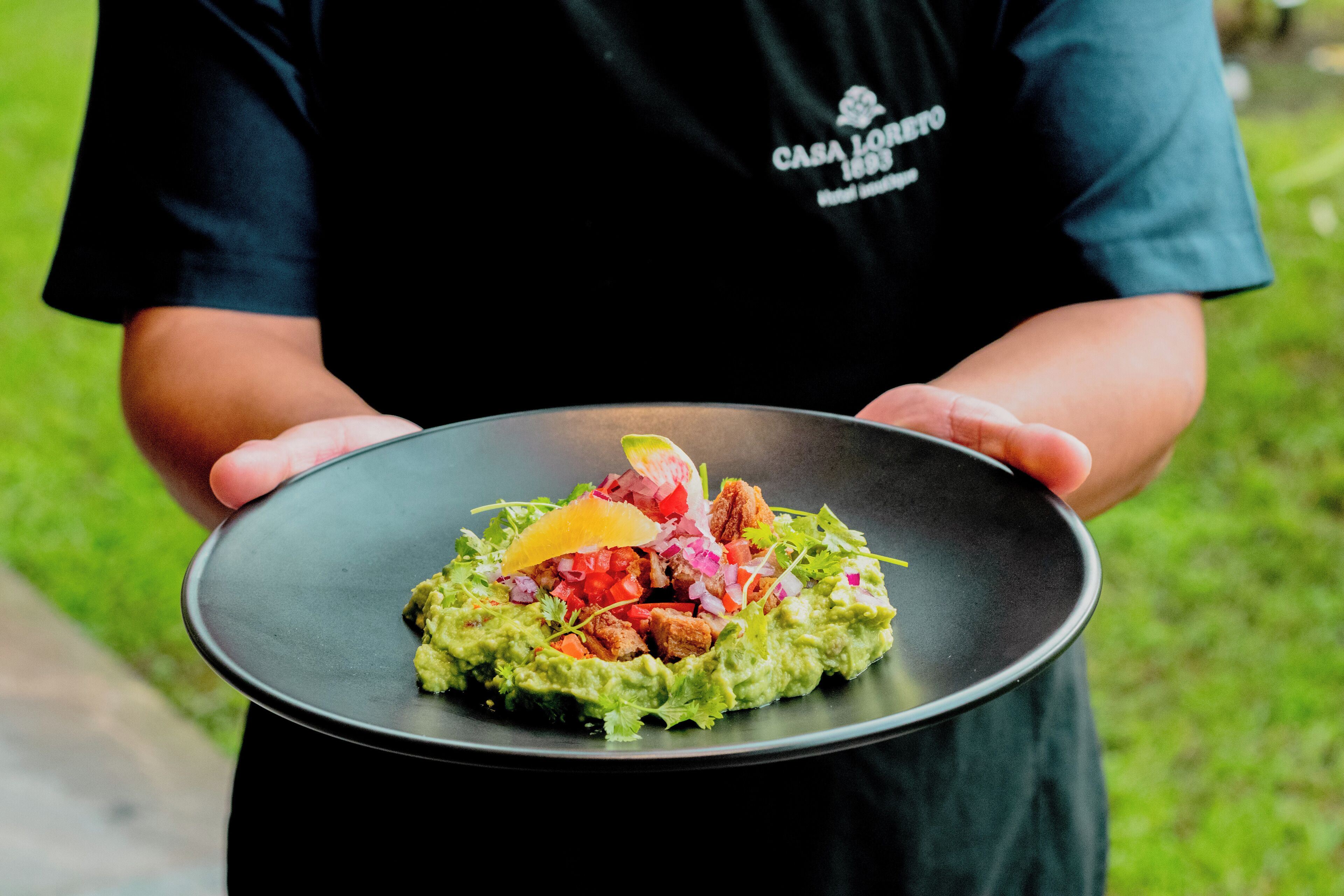 A person holding a black plate with a dish of guacamole, diced tomatoes, onions, and a slice of cheese.