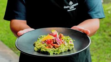 A person holding a black plate with a dish of guacamole, diced tomatoes, onions, and a slice of cheese.