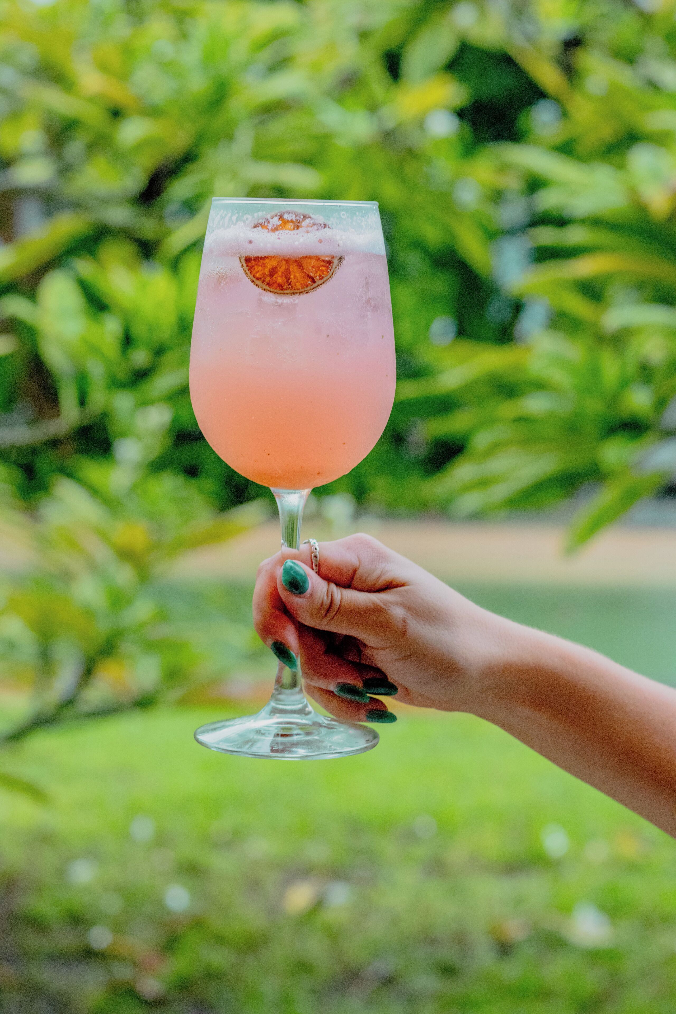 Une main tenant un verre contenant une boisson rose et une tranche de fruit, le tout sur un fond vert flou.