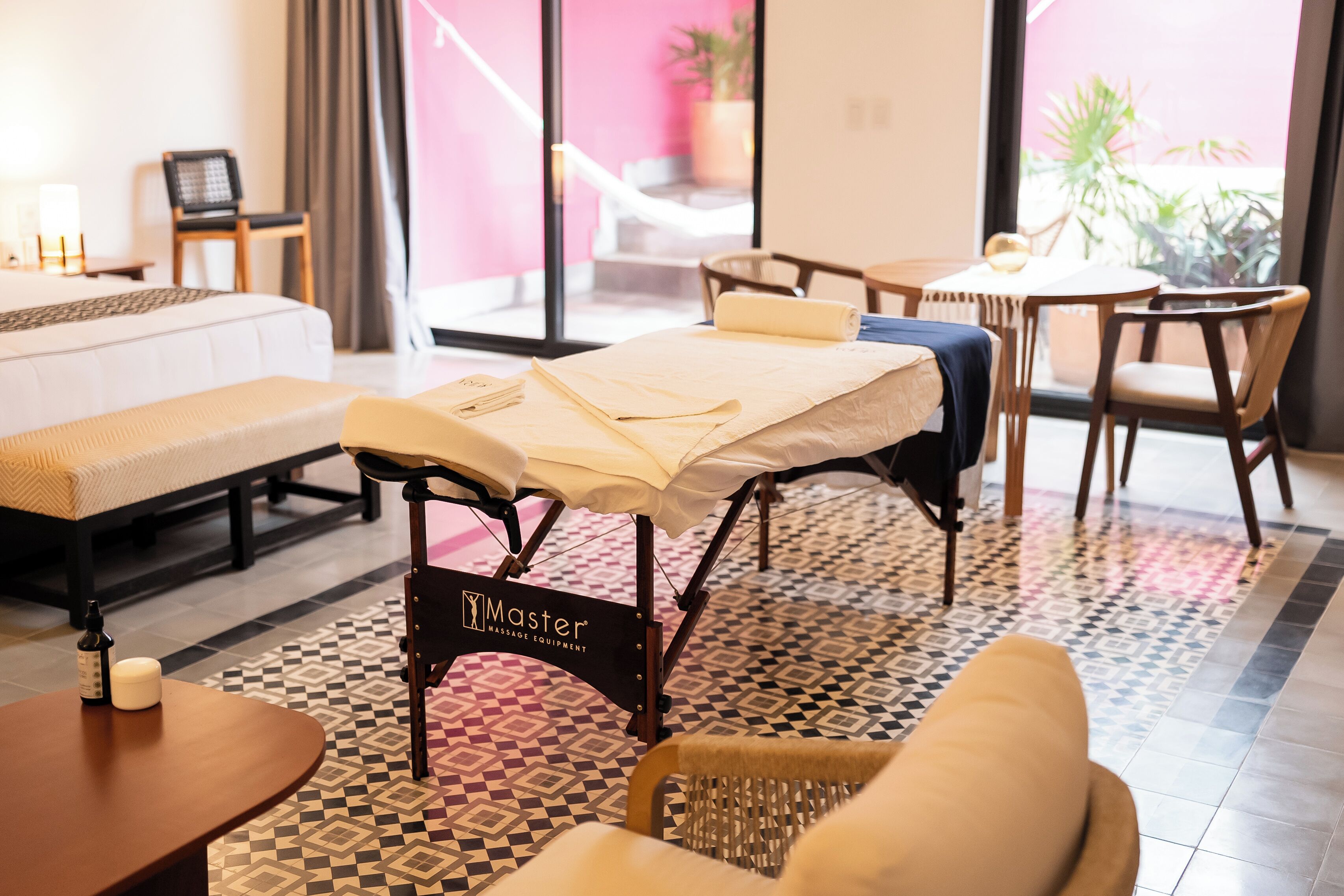 A massage therapy room with a massage table, a chair, a small table with a bottle and a container, and a patterned floor.