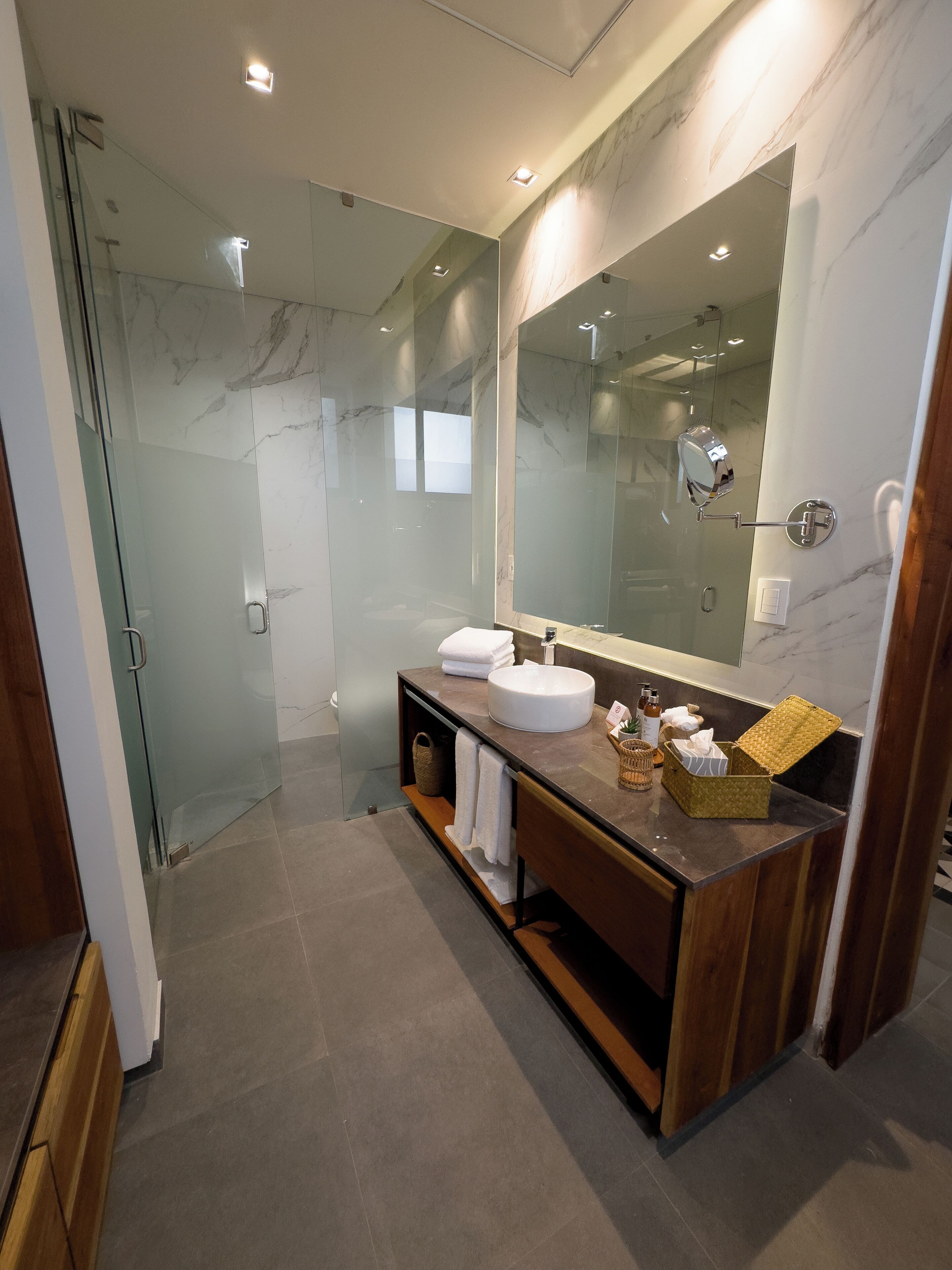 A modern bathroom with a glass-enclosed shower, a marble-tiled wall, and a wooden vanity with a large mirror.