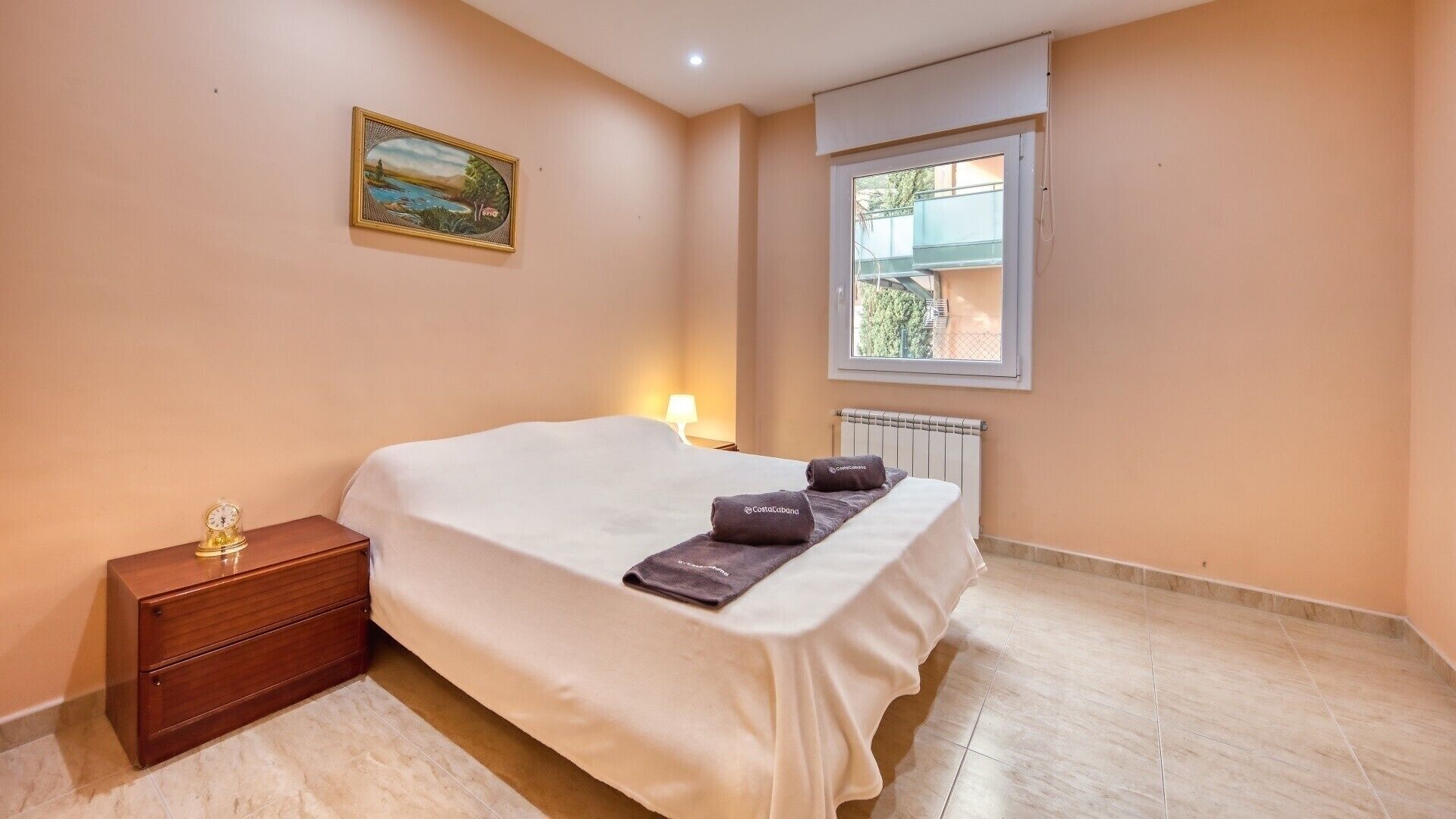 A bedroom with a bed, a nightstand, a lamp, a radiator, a window, and a painting on the wall.