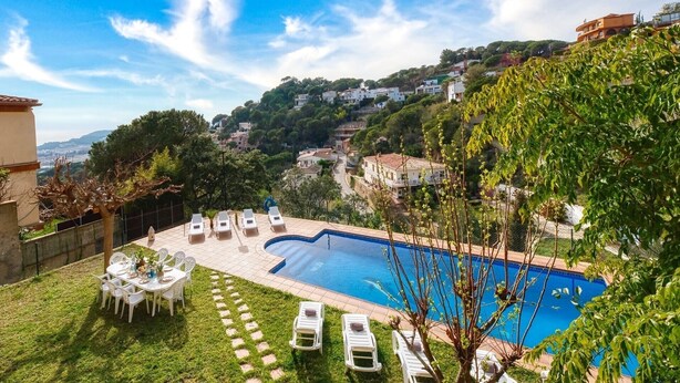 A swimming pool with lounge chairs and a dining table, surrounded by greenery and overlooking a hillside with buildings. - Villa 'Costacabana -  Marysol' with Sea View, Private Pool and Wi-Fi (Lloret de Mar)