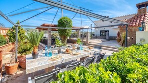 Un patio al aire libre con una mesa lista para comer, rodeado de plantas en macetas y una pérgola.