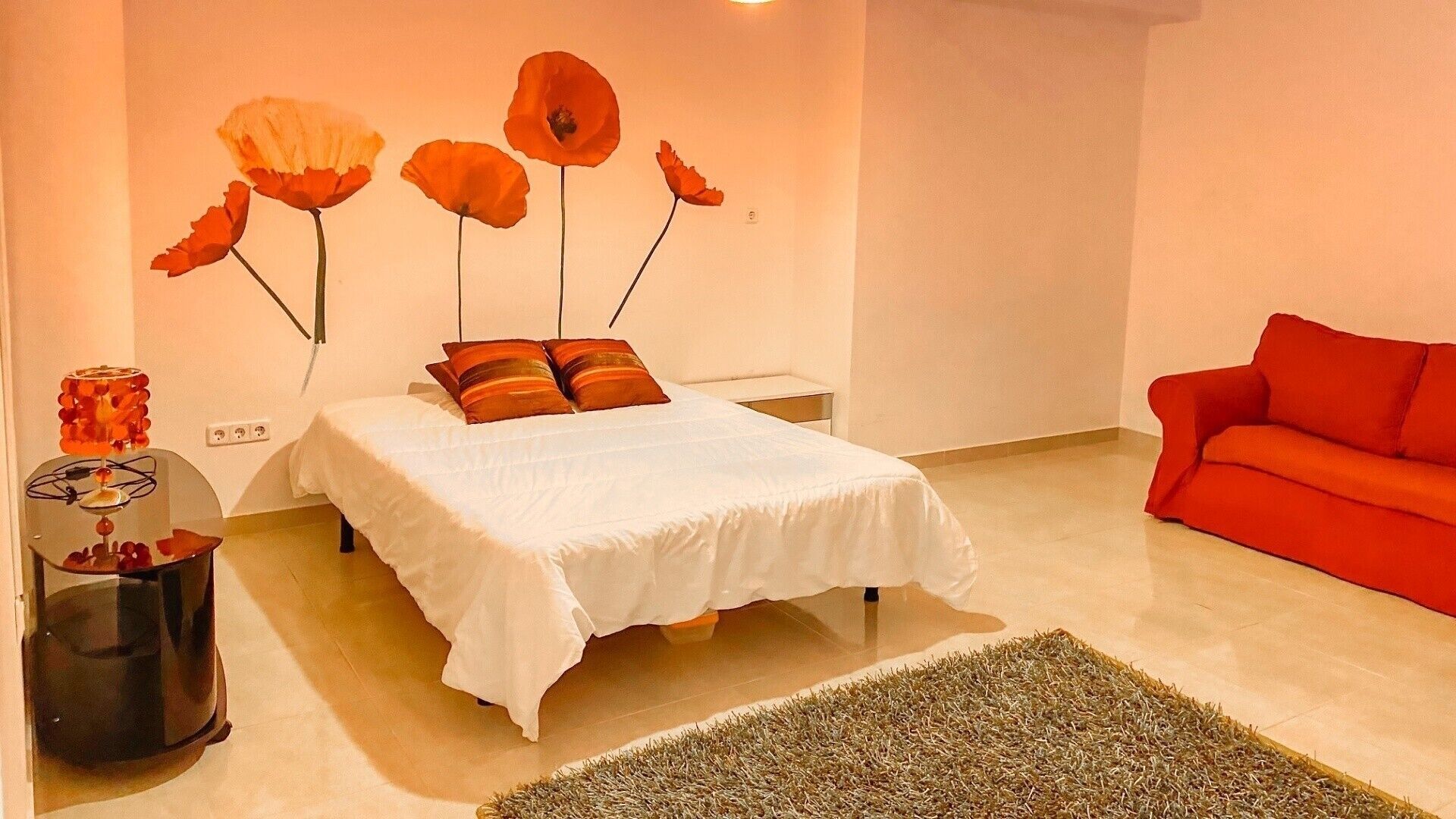 A bedroom with a bed, a red sofa, a wall art piece, and a small table with a lamp.