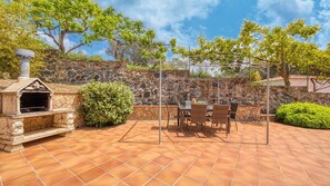 An outdoor patio with a stone barbecue grill, a table with chairs, and a stone wall backdrop. - Villa "Costacabana - Lloret" with Shared Pool, Private Terrace & Wi-Fi (Lloret de Mar)