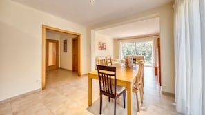 A dining area with a wooden table and chairs, a sofa, and a large window with a view. - Villa "Costacabana - Lloret" with Shared Pool, Private Terrace & Wi-Fi (Lloret de Mar)