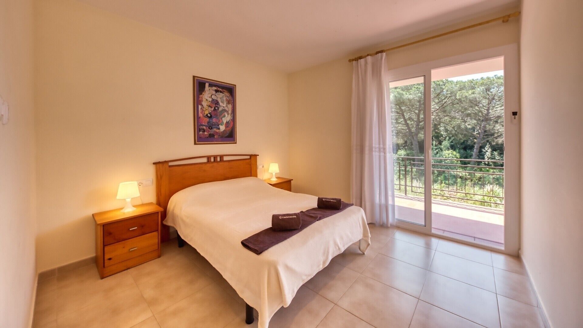 Un dormitorio con cama, dos mesitas de noche con lámparas, un cuadro en la pared y un balcón con vistas a árboles.