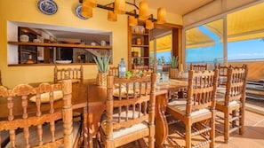A wooden dining table with chairs, a kitchen area with shelves, and a view through large windows.