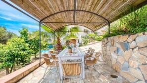 Une terrasse extérieure avec un mur en pierre, un espace salon couvert avec des fauteuils en osier, et une piscine en arrière-plan.
