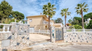 Una entrada con puerta y muros de piedra, un edificio de dos pisos con balcón, y palmeras al fondo.