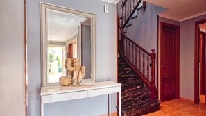 Un couloir avec un grand miroir, une console ornée de divers objets décoratifs, un escalier avec des rampes en bois et une porte.