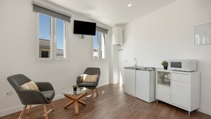 Ein modernes Wohnzimmer mit zwei Sesseln, einem Glastisch, einer weißen Küche mit Mikrowelle und einem wandmontierten Flachbildfernseher.