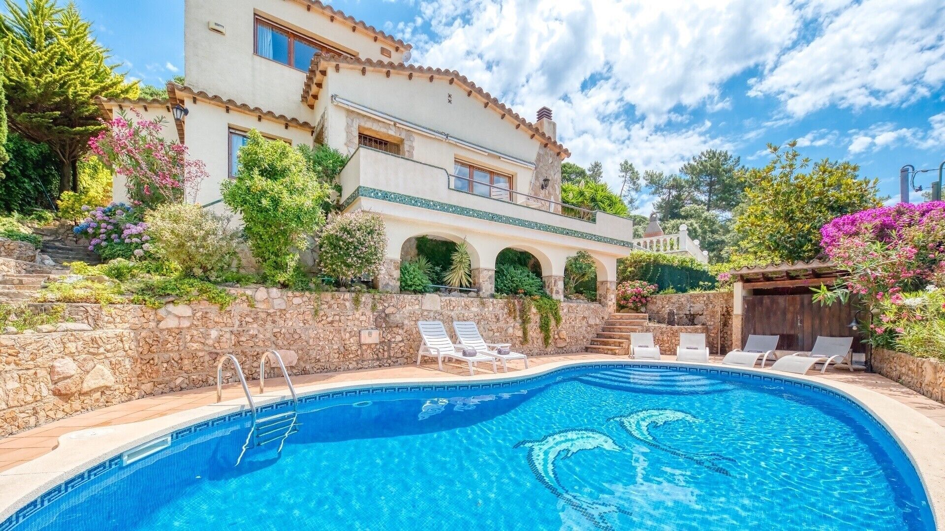 Une piscine bordée d’un mur en pierre, avec des chaises longues et un bâtiment doté d’arcades et d’un balcon.