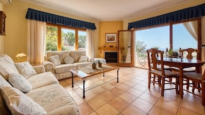 A living room with a sofa, coffee table, and dining table. There is a fireplace and a view through large windows.