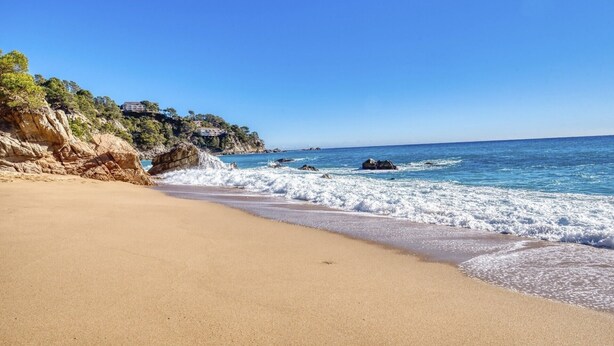 A beach with clear blue water, sandy shore, and rocky cliffs. - Holiday Home "Costacabana - Casa Adora" with Mountain View, Shared Pool & Wi-Fi (Tossa de Mar)
