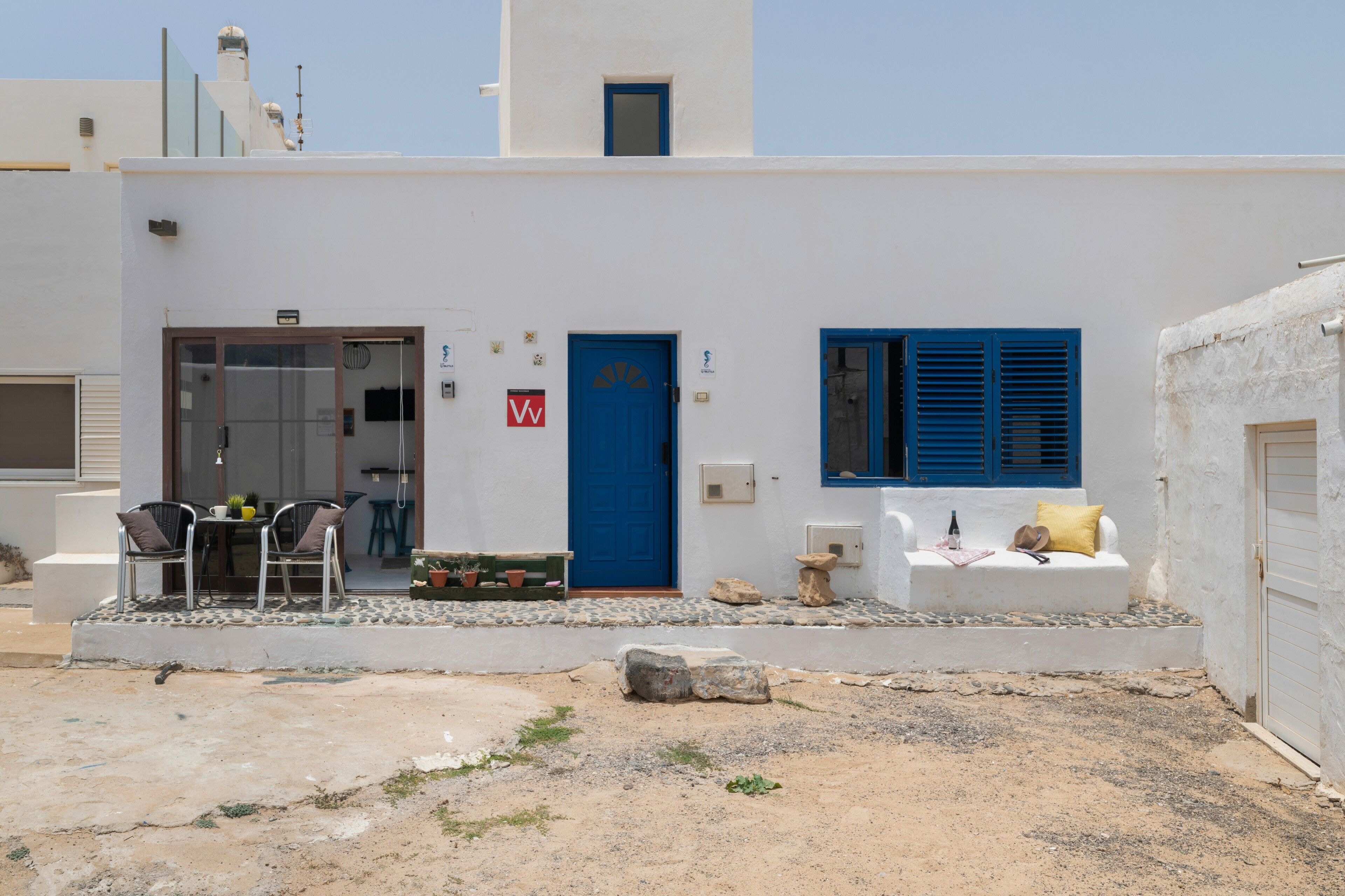 Un edificio blanco con puerta y contraventanas azules, un pequeño espacio al aire libre con mesa y sillas, y un banco de piedra.