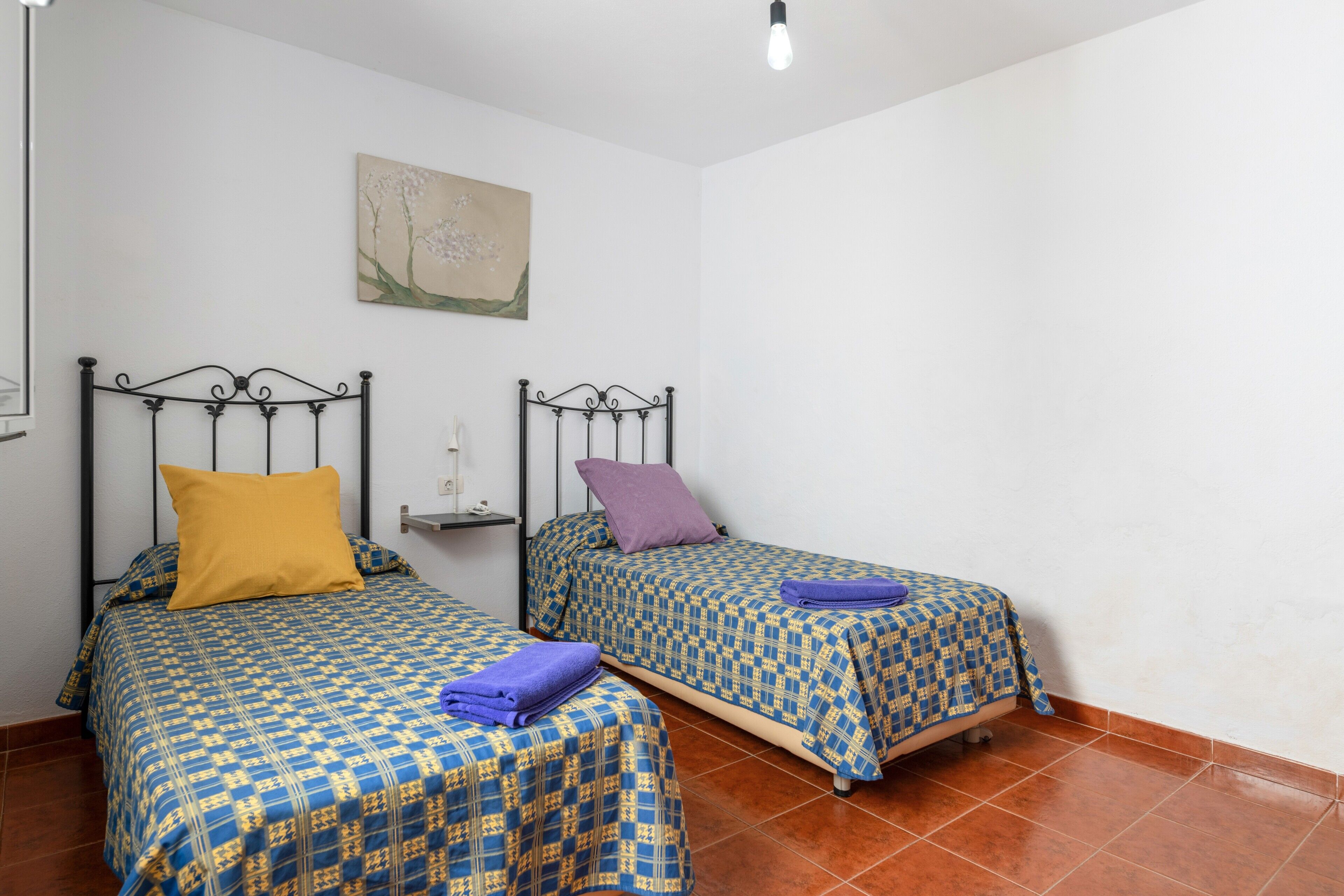 Une chambre avec deux lits, chacun agrémenté d’une couvre-lit et de coussins à motifs, une petite étagère avec une lampe et un tableau au mur.