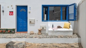 Un bâtiment blanc avec une porte et une fenêtre bleues, un petit espace extérieur aménagé avec des coussins et un chemin en pierre.