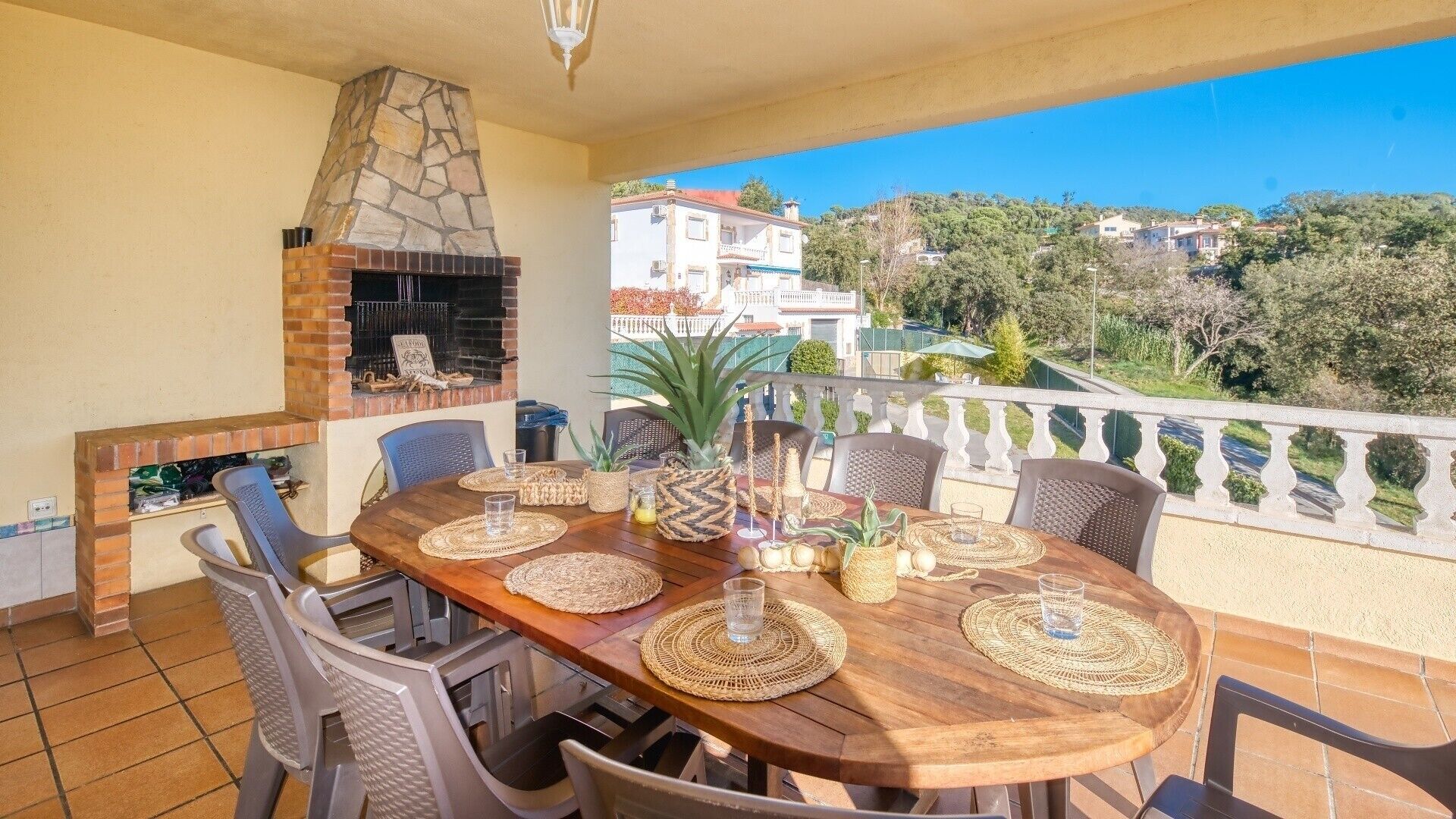 A patio with a stone fireplace, a wooden table set with placemats and glasses, and a view of the surrounding area.