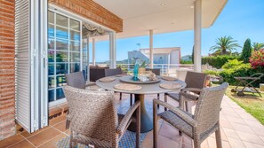 Un espace extérieur abrité, aménagé pour les repas, avec des chaises en osier et une table ronde, de grandes portes coulissantes en verre et une vue sur le paysage environnant.