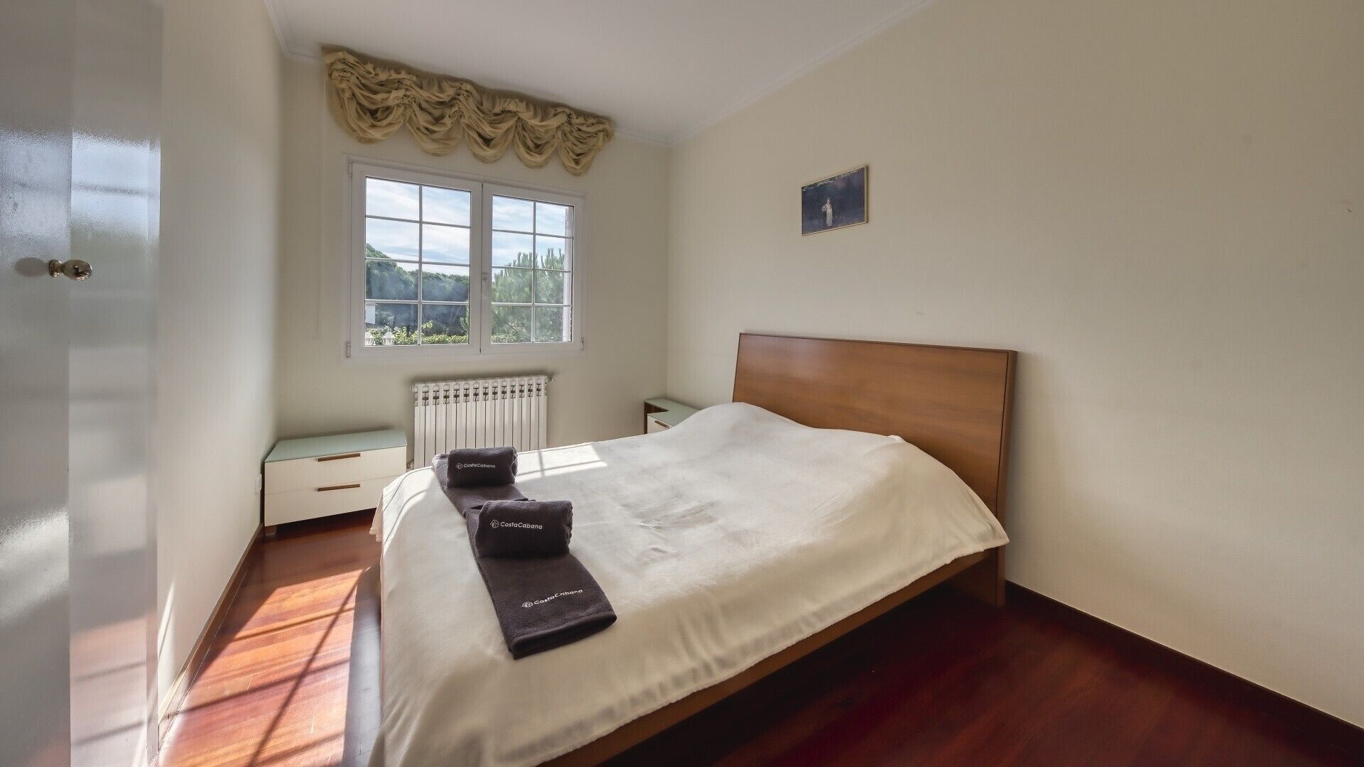 A bedroom with a wooden bed, a radiator, a window with a view, and a small bedside table.