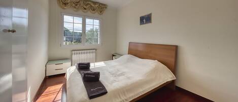 A bedroom with a wooden bed, a radiator, a window with a view, and a small bedside table.