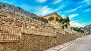 Un mur de pierre surmonté d’une rambarde métallique, un bâtiment doté d’un balcon et un ciel dégagé.