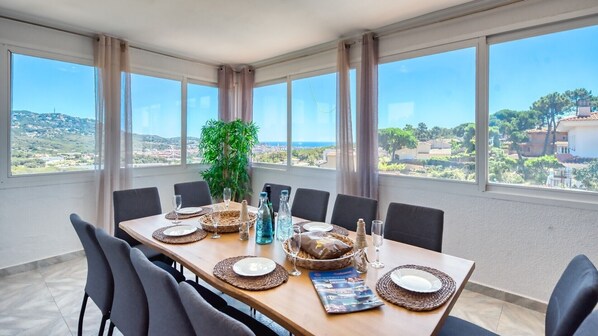 A dining area with a table set for six, large windows, and a view of a coastal landscape.