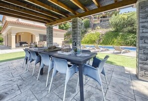 An outdoor patio with a stone table and chairs, a pergola overhead, and a view of a swimming pool and greenery.