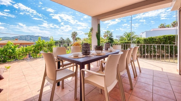 A balcony with a table set for dining, surrounded by chairs, and overlooking a garden and buildings.
