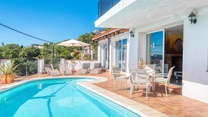 Una piscina con sillas de descanso y una mesa, rodeada por un edificio blanco con ventanas amplias y un balcón.