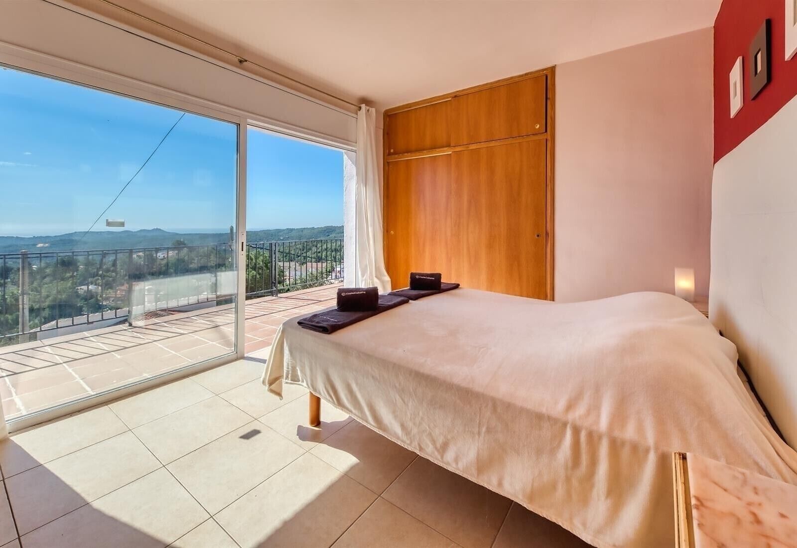 A bedroom with a bed, a wooden wardrobe, a sliding glass door to a balcony, and a view of the landscape.