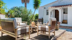 Un área de estar al aire libre con dos sillas de madera y una mesa, rodeada de follaje y un edificio blanco con tejado de tejas.