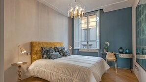 A bedroom with a bed, a round table with a lamp, a chair, a window with a view, and a chandelier. - Apartments Florence - Repubblica Exclusive with Balcony (Florence)