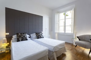 Habitación de hotel con dos camas, un sillón tapizado en gris, una mesita con lámpara y una ventana con vistas a edificios.