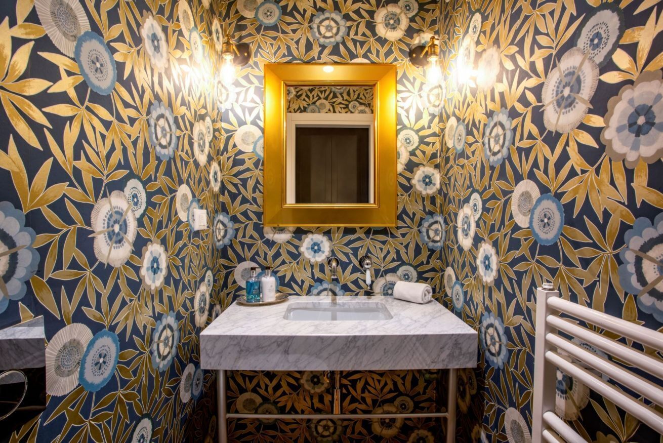 Une salle de bain avec un papier peint à motifs floraux, un lavabo et un miroir encadré d’or.