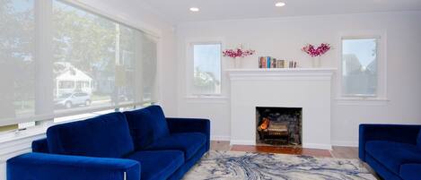 Una sala de estar con chimenea, dos sofás azules, un tapete con estampado de mármol y ventanas amplias.